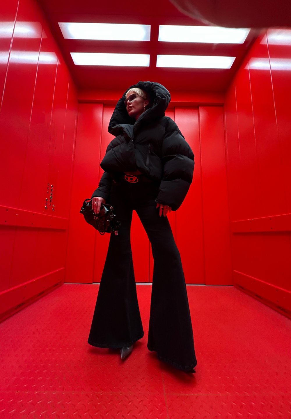 Pessoa com um casaco acolchoado preto e calças largas está numa elevador vermelho, segurando uma mala preta, olhando de lado sob luzes brilhantes no teto.