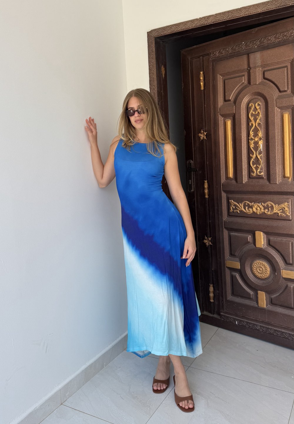 Žena s dlouhými blond vlasy, která má na sobě modré ombré bezrukávové šaty a sluneční brýle, stojí u dřevěných dveří a bílé zdi.
