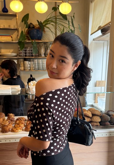 Top marrón de hombros caídos con lunares blancos, combinado con pantalones negros y un pequeño bolso negro. En el fondo se exhiben productos de panadería.