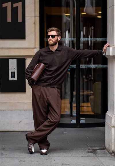 Hombre con ropa marrón oscuro y gafas de sol apoyado en una columna fuera de un edificio, sosteniendo una bolsa marrón bajo un brazo.
