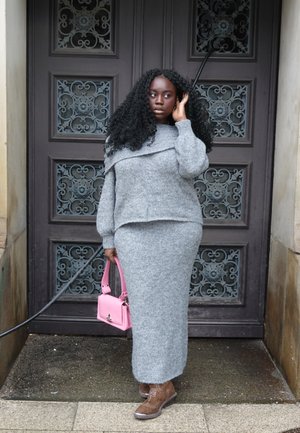 Mujer negra con cabello largo y rizado, vestida con un suéter de punto gris y falda, sosteniendo un bolso rosa, de pie junto a una puerta de madera ornamentada.