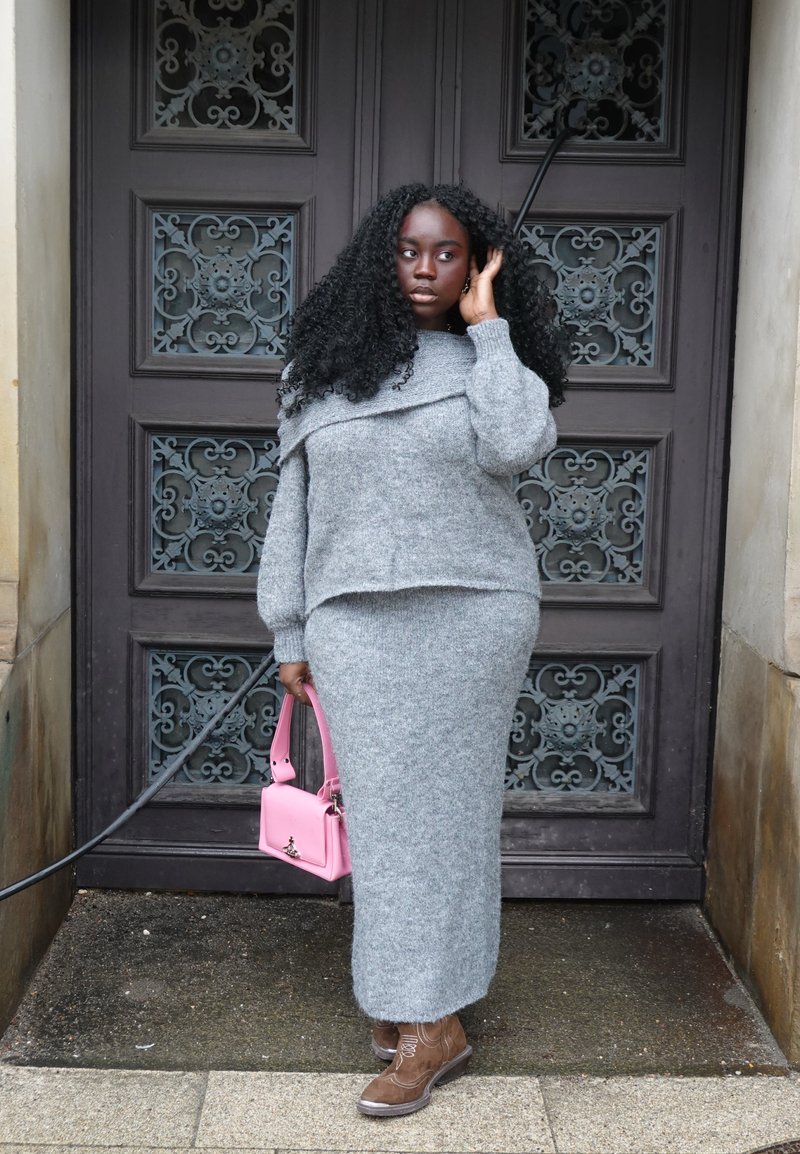 Femme noire aux longs cheveux bouclés portant un pull et une jupe en maille grise, tenant un sac à main rose, debout près d'une porte en bois ornée.