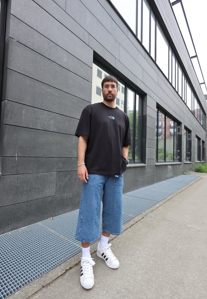 Homme avec des lunettes portant un t-shirt noir, un jean large bleu longueur genoux, des chaussettes blanches et des baskets blanches, debout sur le trottoir près d'un bâtiment gris moderne.