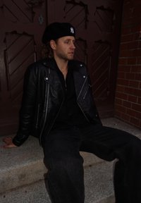 Zwarte leren jas met zilverkleurige hardware, zwart overhemd met knopen, donkere spijkerbroek. Zittend op stenen traptreden tegen een bruine houten deur.