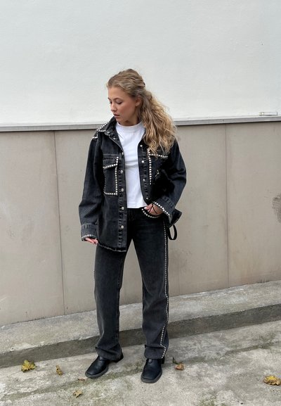 Chaqueta de denim negra con tachuelas plateadas, bolsillos frontales, combinada con pantalones de denim negros a juego. Camiseta blanca debajo. Botines negros.