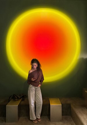 Mujer joven con cabello rizado observa su teléfono bajo una gran luz circular de colores amarillo, naranja y rojo en una pared oscura, cerca de bancos metálicos.