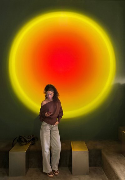 Mujer joven con cabello rizado observa su teléfono bajo una gran luz circular de colores amarillo, naranja y rojo en una pared oscura, cerca de bancos metálicos.