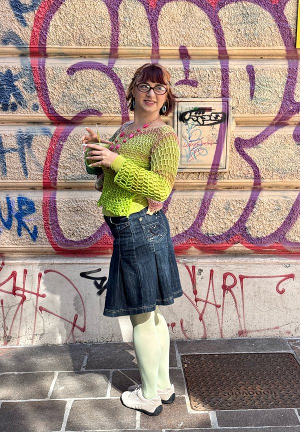Personne avec des lunettes et des cheveux courts portant un pull en maille vert, une jupe en jean et des chaussures blanches, debout tenant une boisson verte devant un mur de graffitis.