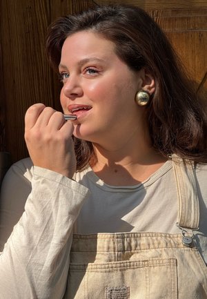 Con lápiz labial en la mano, el modelo se aplica maquillaje. Lleva una camiseta de manga larga en beige claro y un overol beige con una etiqueta de mezclilla y grandes pendientes de oro.