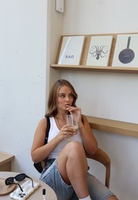 Een vrouw in een witte tanktop en denim shorts drinkt uit een glas. Ze zit op een houten stoel met een minimalistisch ontwerp.