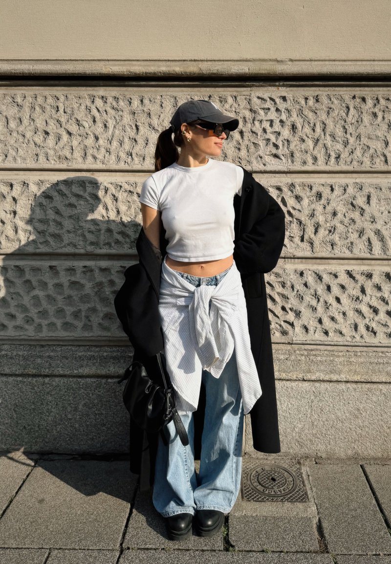 Femme portant une casquette noire, des lunettes de soleil, un t-shirt blanc court, un manteau noir, un jean clair avec une chemise blanche nouée autour de la taille, debout contre un mur en pierre texturé.