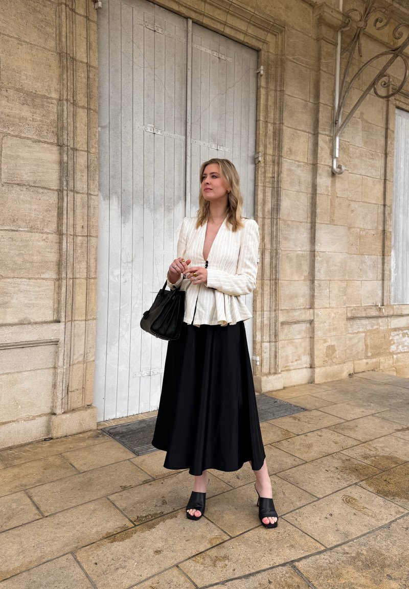 Femme en blazer blanc et jupe noire tenant un sac à main noir, debout sur un pavé en pierre devant des portes en bois beige fermées.