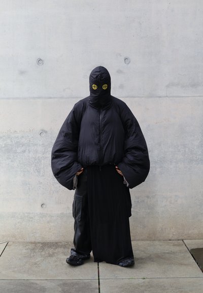 Chaqueta de plumas negra con mangas sobredimensionadas, combinada con una larga falda negra. Diseño con capucha que presenta ojos amarillos en forma de globo. Tejido texturizado.