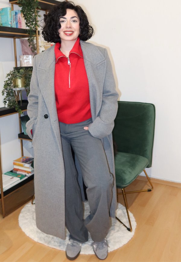 Palton lung gri peste un pulover roșu cu fermoar și pantaloni gri. Pantofi de culoare gri deschis. Scaun verde și rafturi pline cu obiecte în fundal.