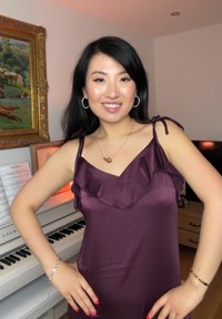 Γυναίκα που χαμογελά, με τα χέρια στη μέση, φορώντας ένα μωβ φόρεμα, στέκεται δίπλα σε ένα λευκό πιάνο με παρτιτούρες και κορνιζαρισμένους πίνακες.