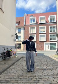 Una persona está de pie sobre adoquines, vistiendo una chaqueta negra con cremallera, una camisa blanca con cuello, pantalones grises de pierna ancha y zapatos negros, ajustándose los auriculares.