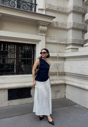 Top azul marino de un solo hombro combinado con una falda maxi larga de encaje blanco transparente. Tacones negros de punta afilada y gafas de sol negras completan el conjunto.