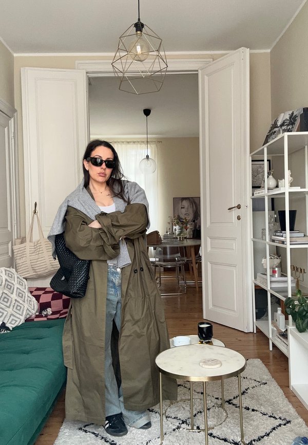 Femme portant des lunettes de soleil, manteau vert olive avec un pull gris posé sur les épaules, debout dans un salon moderne aux murs blancs et à la décoration épurée.