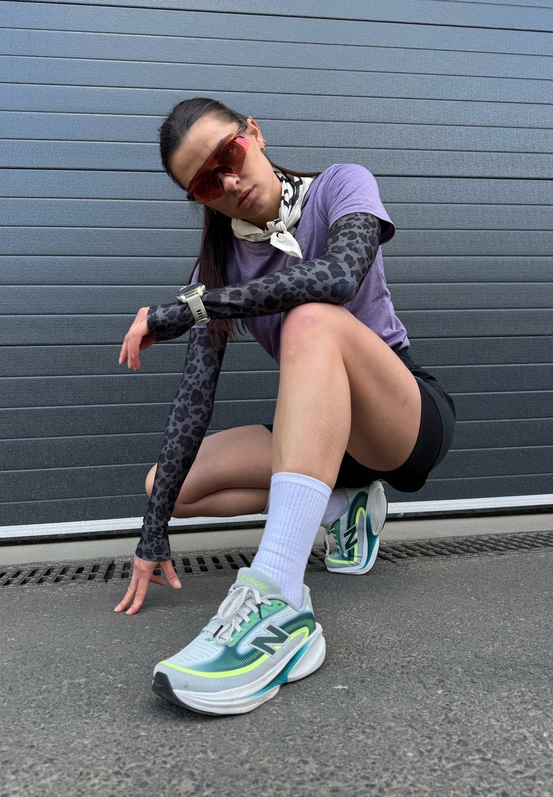 Jeune femme en tenue de sport accroupie sur le trottoir, portant des lunettes de soleil, un t-shirt violet, des manchons à motifs, des chaussettes blanches et des chaussures de course vertes.