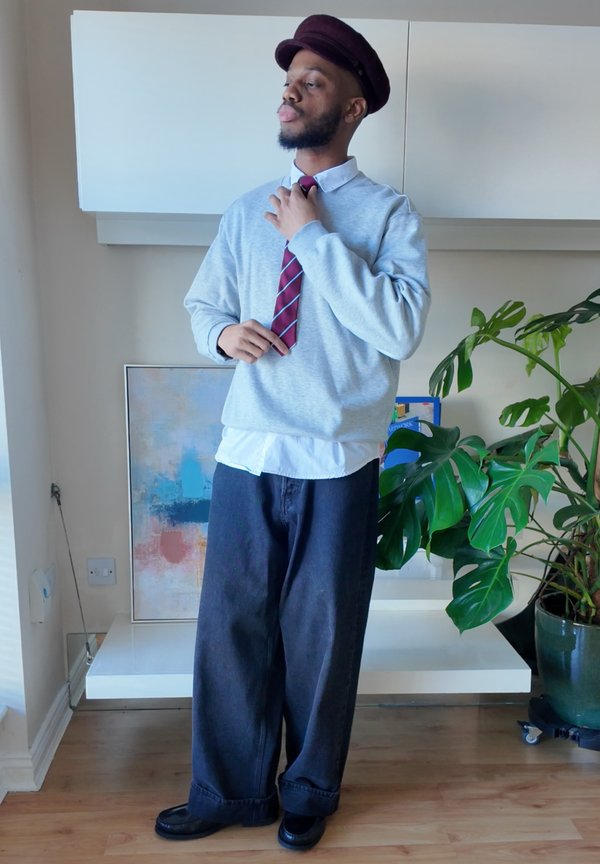 Mann iført en burgunderfarget hatt, lys grå genser, hvit skjorte, stripete rød slips og vide svarte bukser som justerer slipsen sin innendørs nær en stor grønn plante.