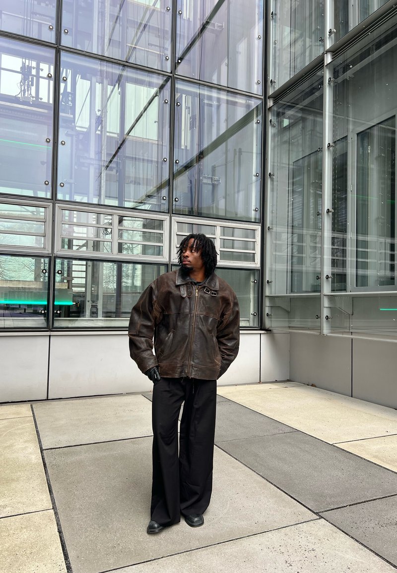 Homme aux dreadlocks portant une veste en cuir marron, un pantalon noir à jambes larges et des gants, debout près d'une grande façade en verre de bâtiment.