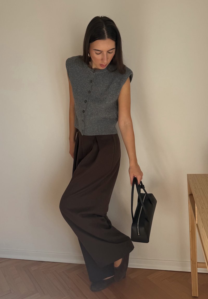 Grey knitted sleeveless top with front buttons, paired with brown wide-leg trousers. Black handbag with zipper, high-heeled shoes. Wood flooring.
