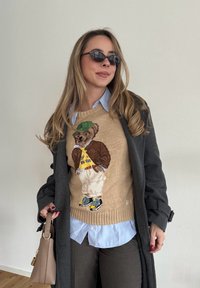 Pull beige avec un motif d'ours portant une casquette verte et une chemise jaune, superposé à une chemise à col bleu clair, avec un manteau gris foncé et un pantalon marron.