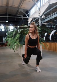 Mustad spordirinnahoidja ja retuusid naisel, kes teeb väljaasteid hantlitega. Sisejõusaal suurte taimede ja marmorist detailidega taustal.