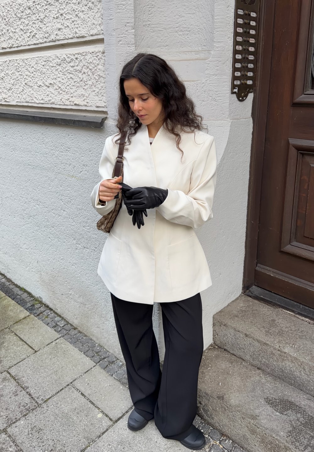 White tailored blazer, black wide-leg trousers, black gloves, and a brown patterned bag. Standing against a textured wall, focusing on accessories.