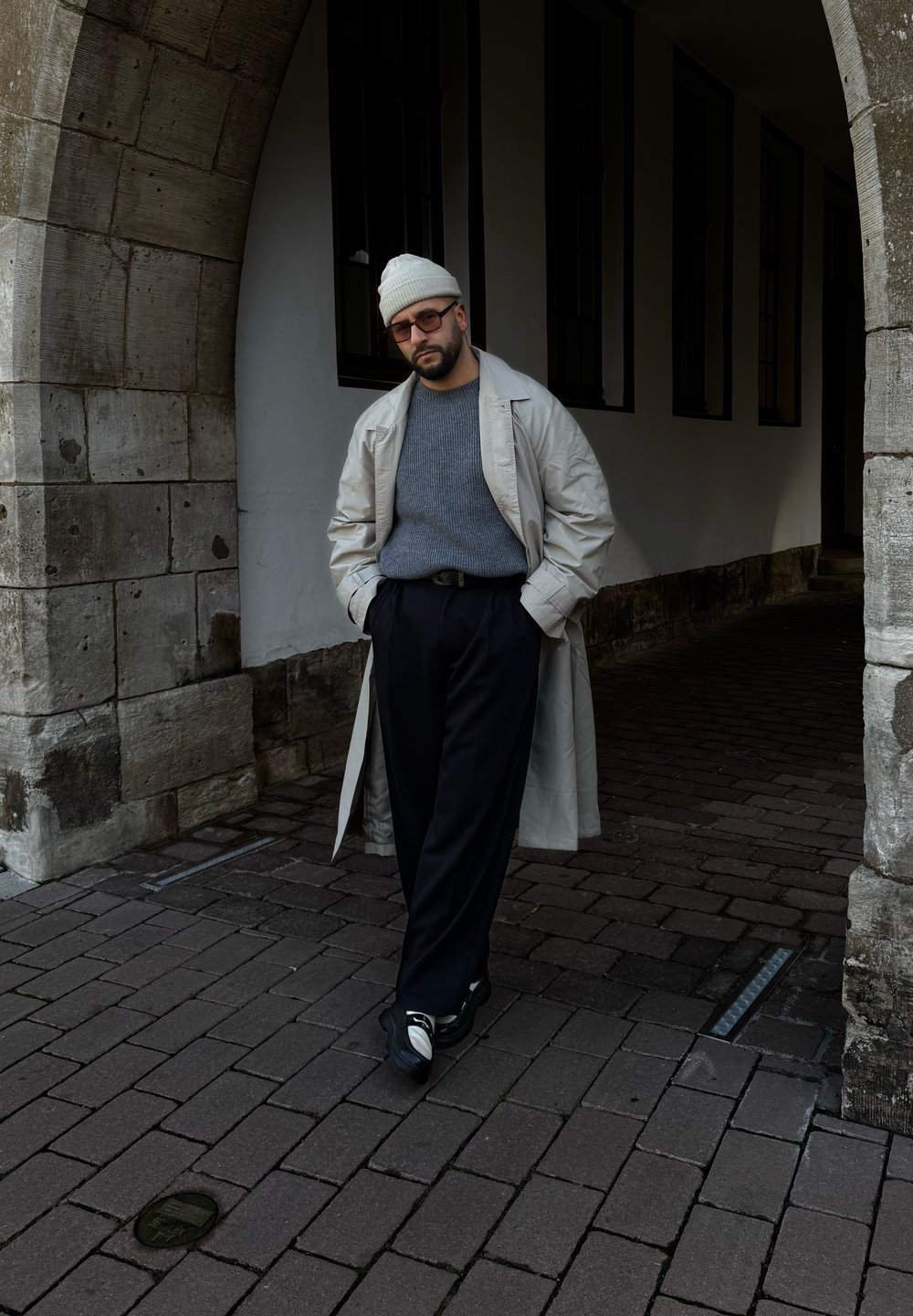 Ανοιχτό μπεζ παλτό πάνω από γκρι πουλόβερ, μαύρο παντελόνι και μαύρο-άσπρα sneakers; στέκεται μπροστά από μια πέτρινη αψίδα.