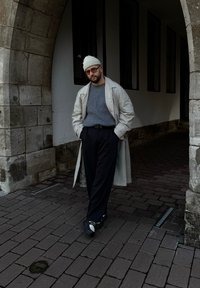 Ανοιχτό μπεζ παλτό πάνω από γκρι πουλόβερ, μαύρο παντελόνι και μαύρο-άσπρα sneakers; στέκεται μπροστά από μια πέτρινη αψίδα.