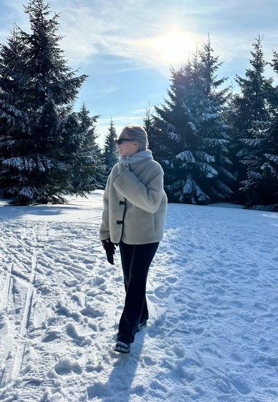 Chaqueta de forro polar gris claro con botones negros, combinada con pantalones negros. Suelo cubierto de nieve y árboles perennes en un entorno exterior soleado.