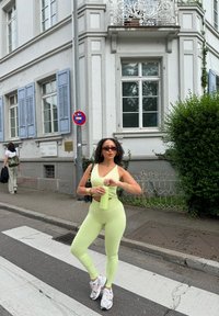 Lys grønn treningsdrakt med en kort topp og tettsittende leggings. Holder en grønn vannflaske, iført hvite og sølvfargede joggesko.