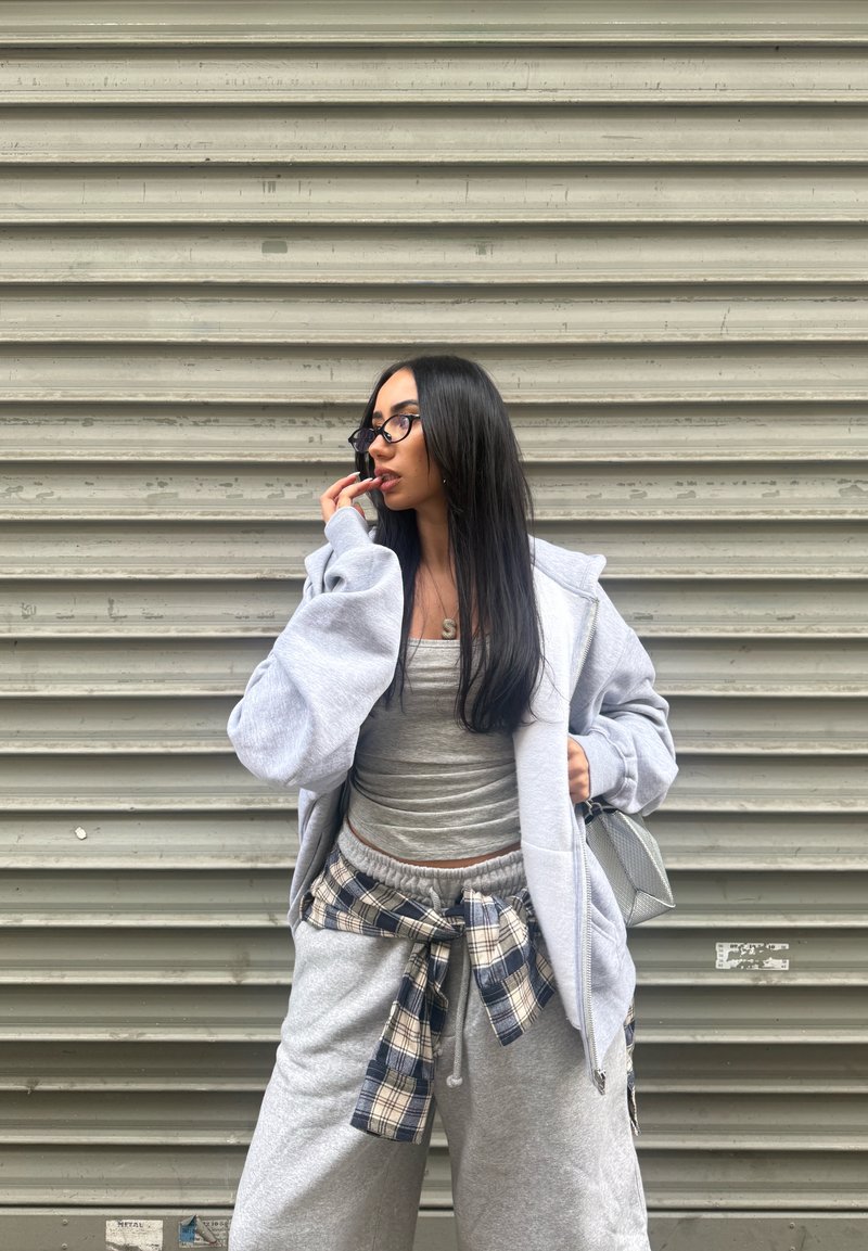 Jeune femme aux longs cheveux noirs et lunettes, vêtue de vêtements casual gris, debout contre un mur métallique ondulé, main près de la bouche.