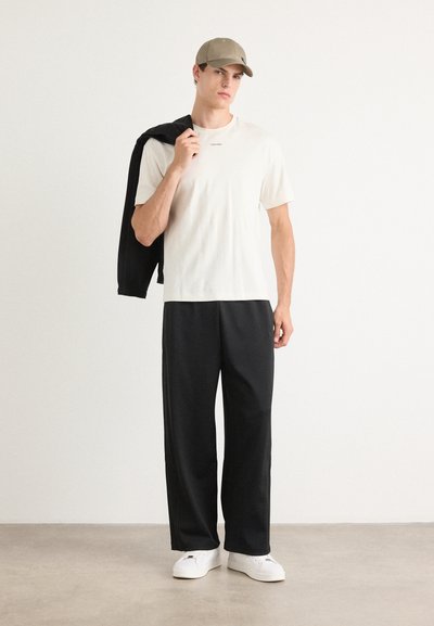 Pantalones negros holgados, camiseta beige clara con logotipo y una gorra beige; de pie con una chaqueta negra sobre un hombro.