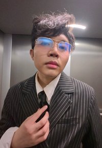 Jeune personne portant des lunettes ajuste une cravate noire, vêtue d'un costume à fines rayures et d'une chemise blanche, à l'intérieur d'un ascenseur métallique.