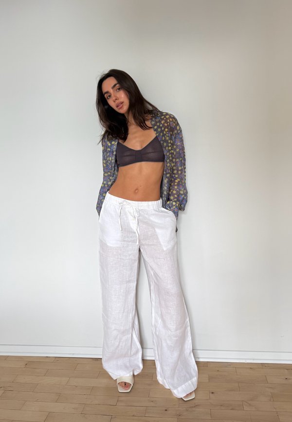 Kobieta stojąca wewnątrz, ubrana w ciemny bralette, kwiatowy przezroczysty top z długim rękawem, luźne białe lniane spodnie i odkryte szpilki na tle gładkiej ściany.