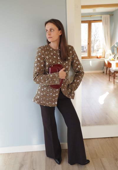Chaqueta marrón adornada con patrones florales, combinada con pantalones negros acampanados y un bolso de mano burdeos; textura suave y diseño ajustado.