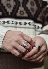 Hands adorned with two silver rings featuring intricate designs, set against a background of a brown and cream patterned sweater.