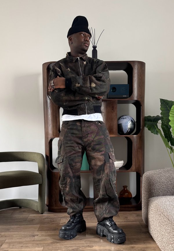 Homme en veste et pantalon camouflage, bonnet noir et bottes noires épaisses, debout les bras croisés dans un salon moderne avec des étagères en bois.