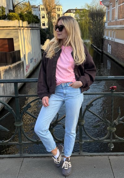 Una mujer está de pie en un puente, vistiendo una chaqueta bomber marrón oscura, una camiseta rosa, jeans azul claro y zapatillas grises con cordones blancos.