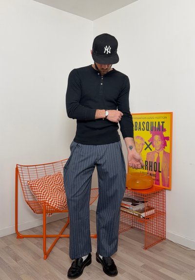 Hombre con camisa negra de manga larga y pantalones de rayas ajustando la manga, de pie junto a una silla de alambre naranja y una mesa auxiliar con un póster de Basquiat.