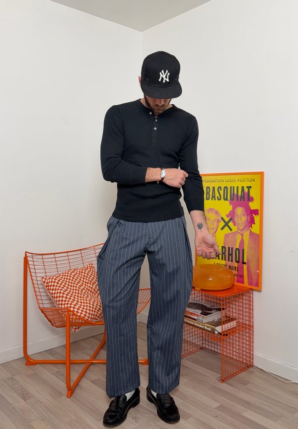 Mann i svart langermet skjorte og bukser med tynne striper justerer ermet, står ved en oransje trådstol og et sidebord med Basquiat-plakat.