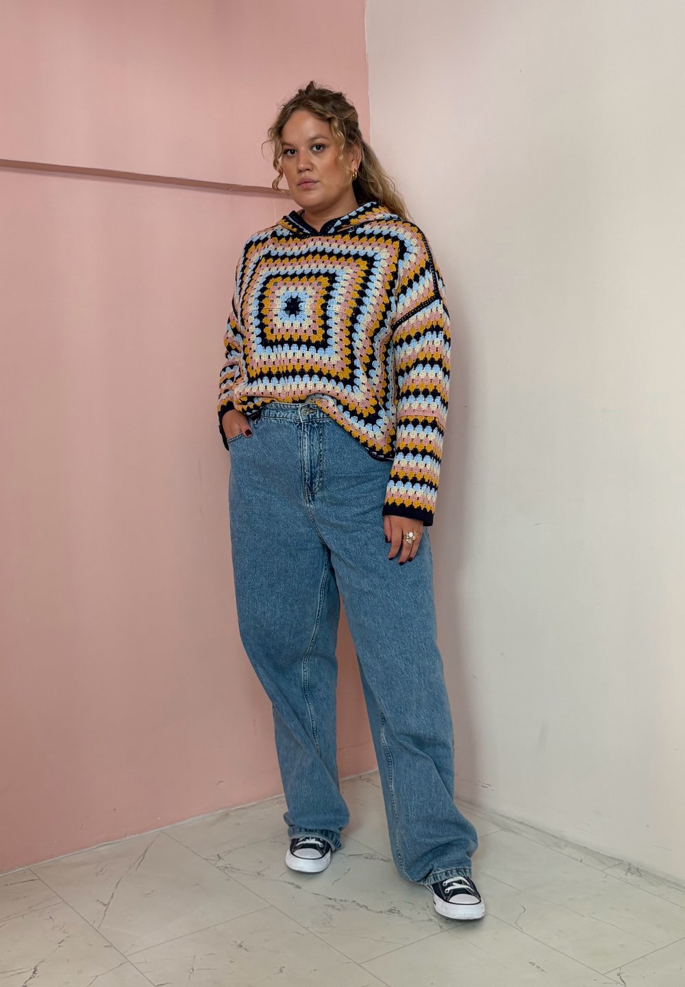 Moletom de croché multicolorido com padrões geométricos, apresentando tons de amarelo, azul e preto. Usado com jeans largos de tom azul claro e sapatilhas pretas.