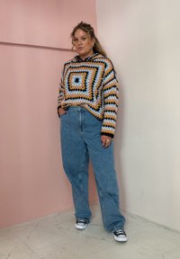 Moletom de croché multicolorido com padrões geométricos, apresentando tons de amarelo, azul e preto. Usado com jeans largos de tom azul claro e sapatilhas pretas.