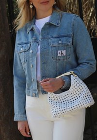 Veste en denim bleu clair avec un patch logo, haut blanc et sac en osier blanc avec des accents dorés. Le modèle tient le sac et porte des boucles d'oreilles.