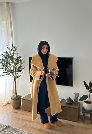 Abrigo largo de lana beige con cuello ancho, combinado con un suéter de cuello alto negro y jeans oscuros. Sosteniendo una revista, de pie cerca de una planta en maceta.