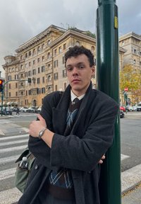 Vīrietis atbalstās pret zaļu stabu, valkājot tumšu mēteli pār brūnu un zilu raibu džemperi ar sudraba pulksteni uz plaukstas.