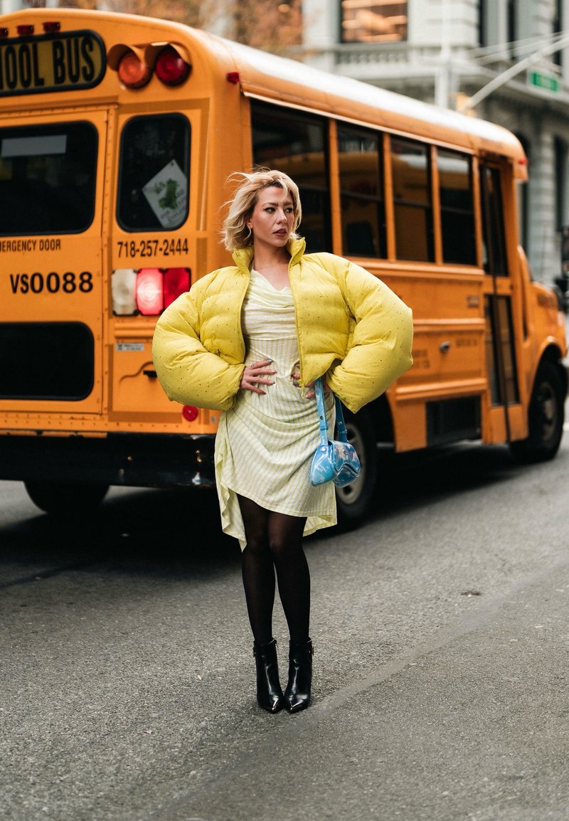 Donna con giacca imbottita gialla e abito che sta con sicurezza per strada davanti a un autobus scolastico giallo, tenendo una piccola borsa blu.