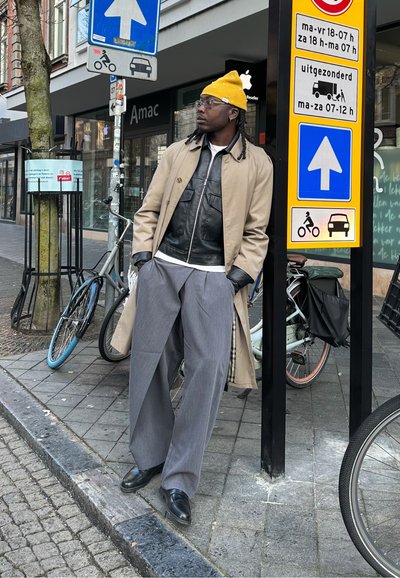 Hombre con gorro amarillo, abrigo beige y pantalones grises apoyado en un poste de señalización cerca de bicicletas estacionadas en una acera de la ciudad.